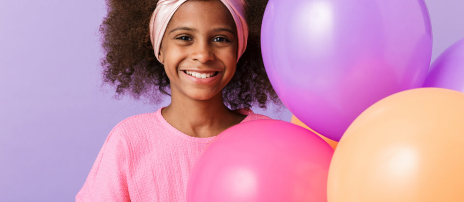 Smiling girl with curly hair and a headband holds pink, purple, and orange balloons against a purple background.