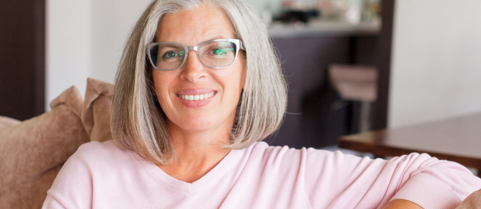 A smiling woman with gray hair and glasses sitting on a couch, wearing a pink sweater, in a living room setting.