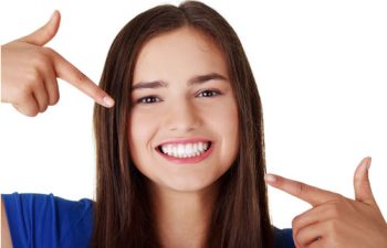 A woman with long hair smiles widely, pointing to her teeth with both index fingers.