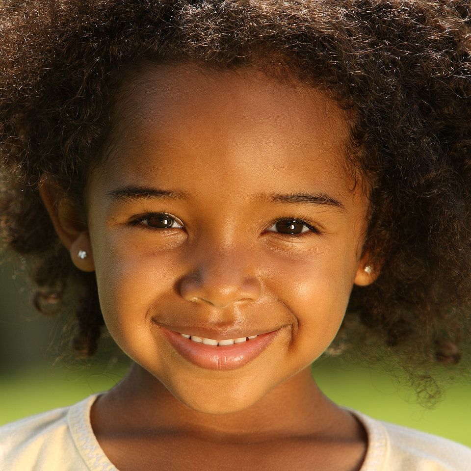 Young child smiling outdoors, with curly hair and wearing small earrings.