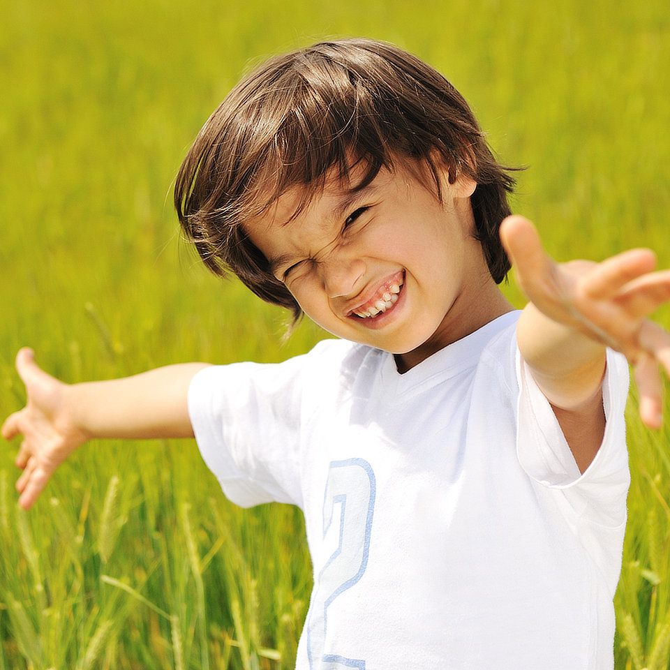 Child in a white shirt stands in a grassy field, smiling broadly with arms outstretched.