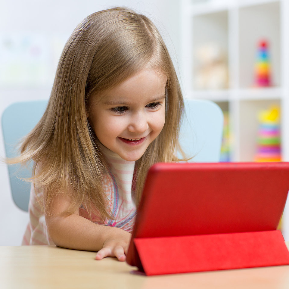 Child smiling while using a red tablet, sitting at a table in a room with colorful toys in the background.