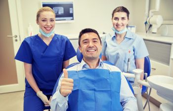 A man in a dental chair gives a thumbs up, flanked by two smiling dental professionals wearing scrubs and face masks, in a dental office setting.