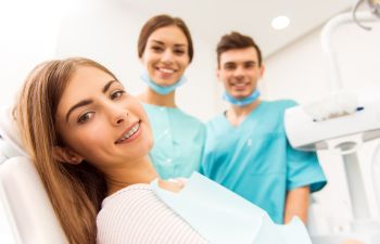 A woman with braces sits in a dental chair, smiling. Two dental professionals in scrubs and masks stand behind her, smiling.