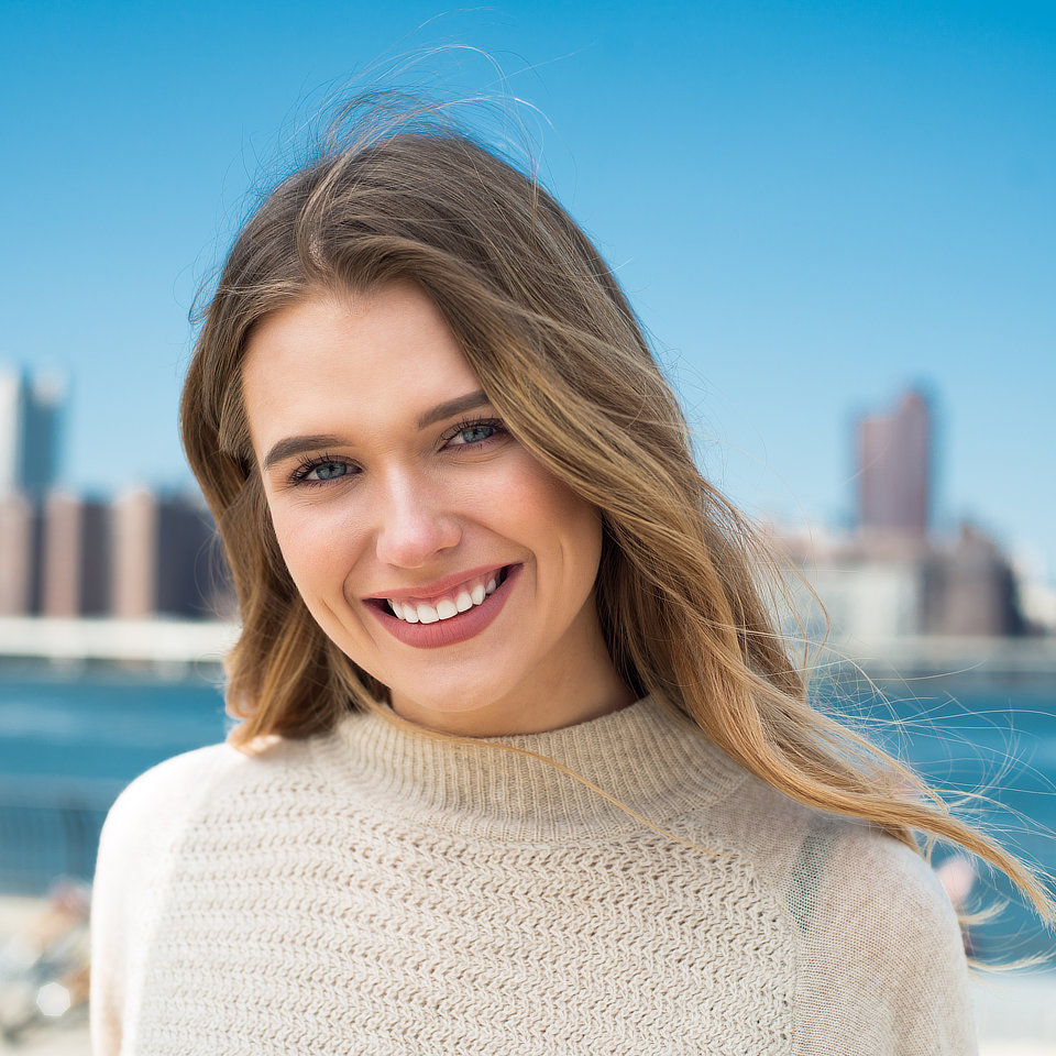 A person with long hair smiles while standing outdoors. A city skyline and body of water are visible in the background.