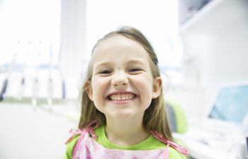 A smiling girl in a green shirt with a pink apron stands indoors, with blurred office equipment in the background.