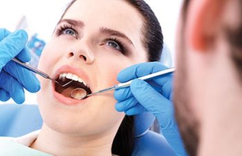 A dentist examines a woman's teeth using dental tools. She is seated in a dental chair, and both are wearing gloves.