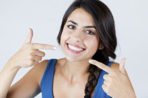 Woman smiling and pointing at her teeth with both hands, wearing a blue top.