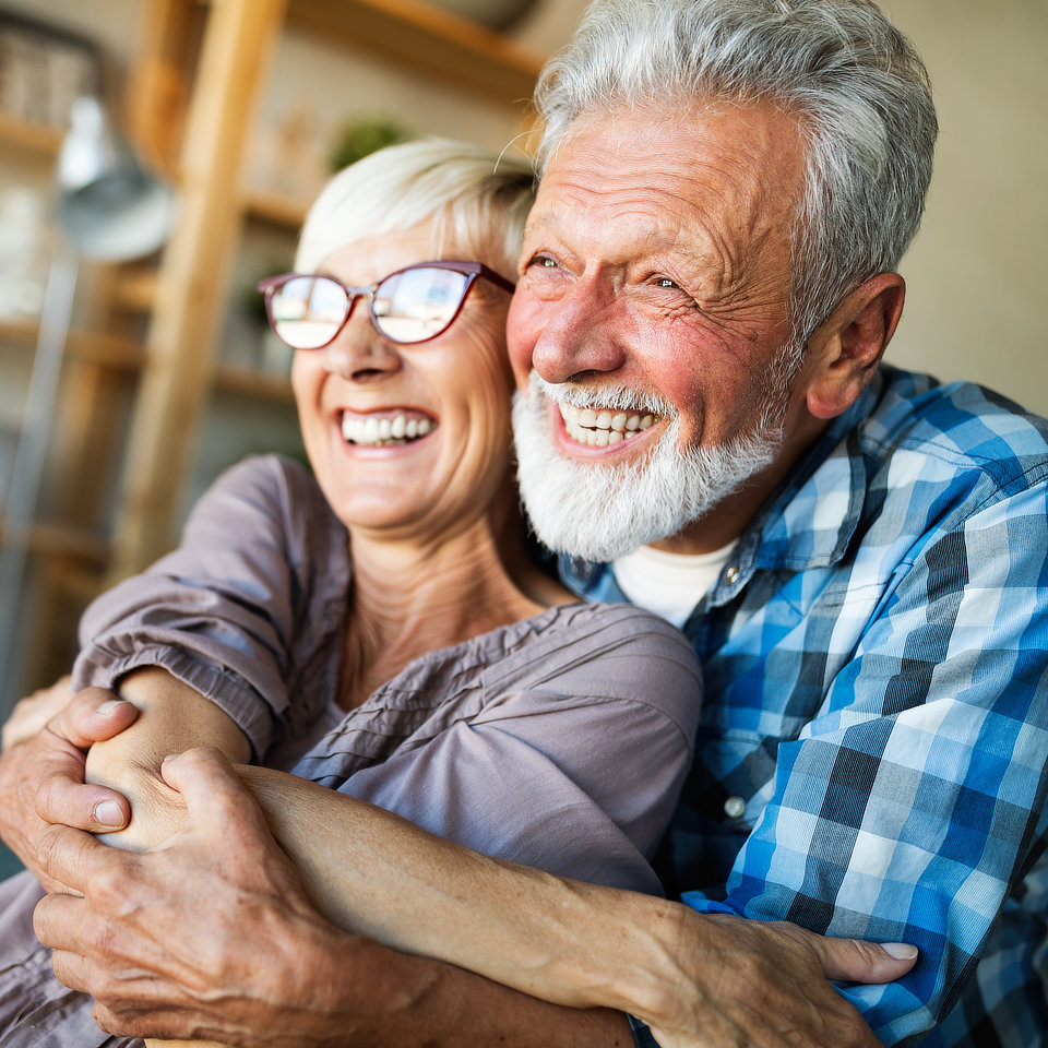 Older couple smiling and embracing each other in a warmly lit room. The man is wearing a plaid shirt, and the woman is wearing glasses and a blouse. They appear happy and content.