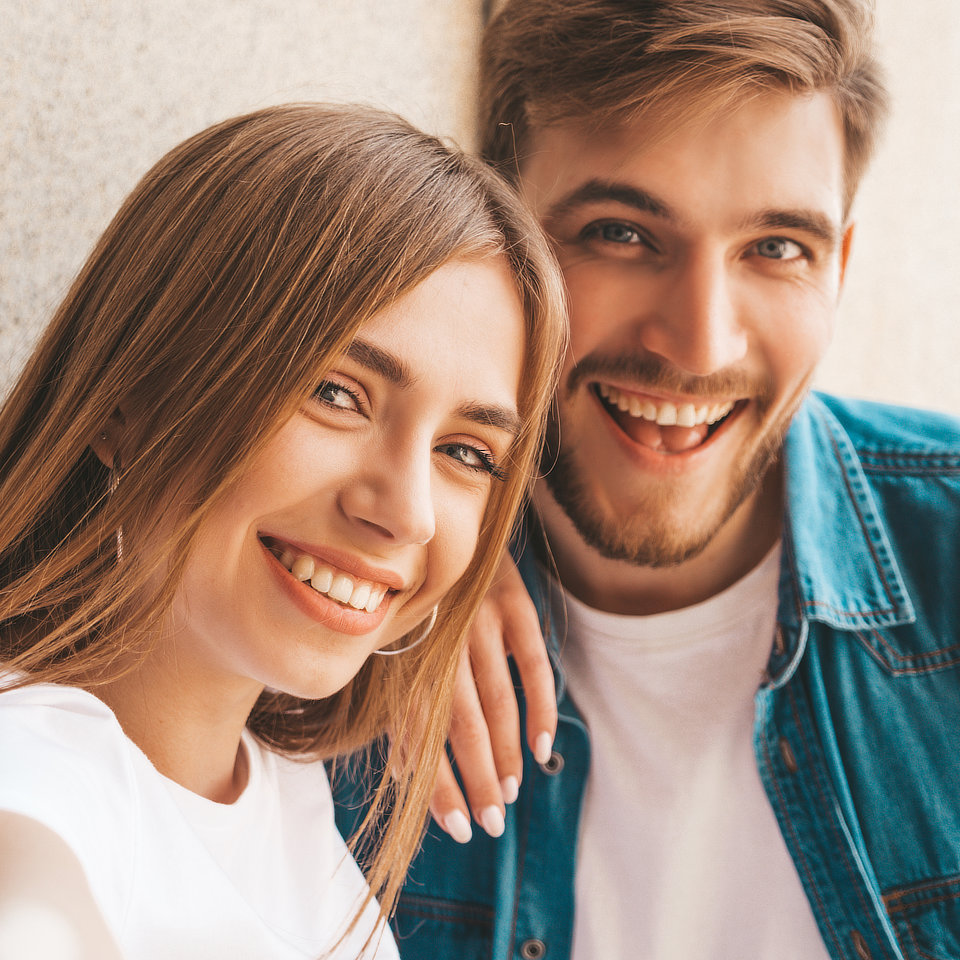 Two people smiling, one with long hair in a white shirt, the other with a beard in a denim shirt, taking a selfie indoors.