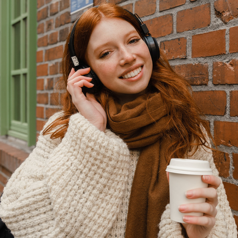 Person with red hair, wearing headphones, holds a coffee cup and smiles while standing against a brick wall.