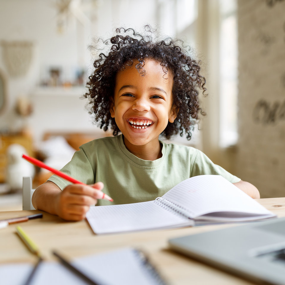 Child with curly hair smiles while sitting at a table with a notebook and holding a red pencil, in a bright room.