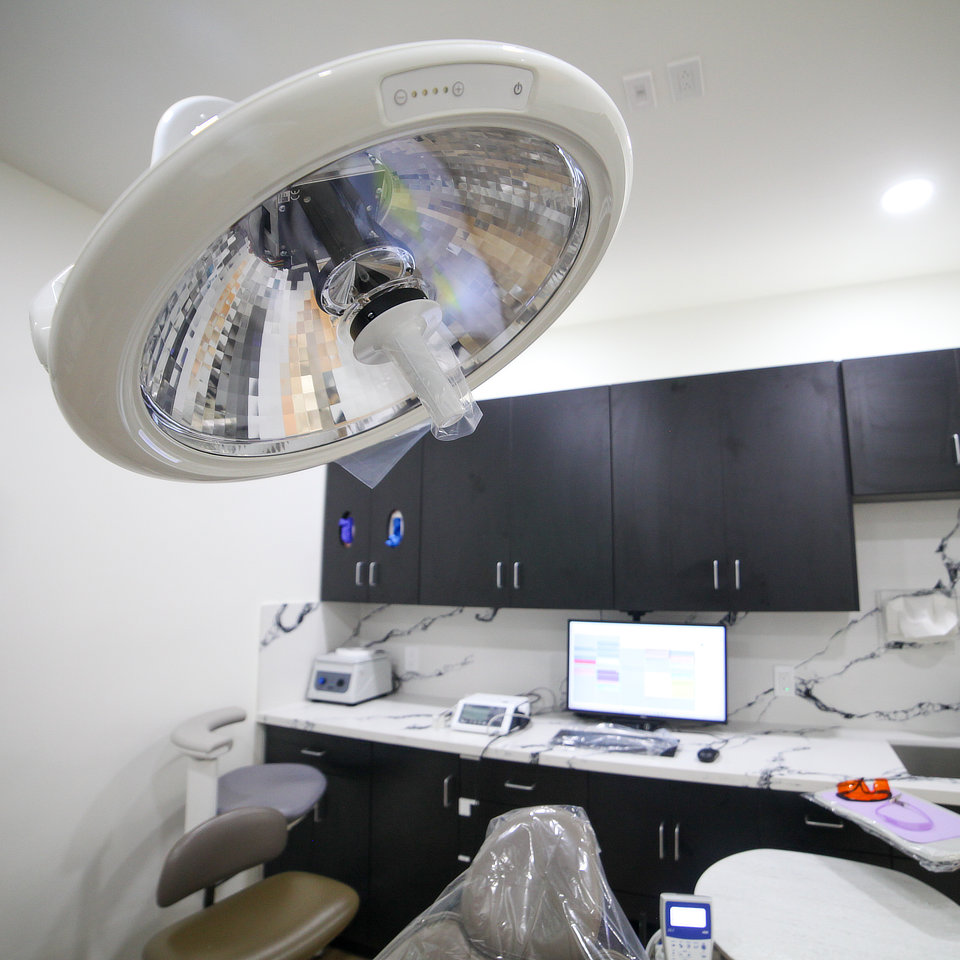 Dental office with chair, overhead light, and computer. Black cabinets and countertop with dental tools. White walls and marble backsplash visible.