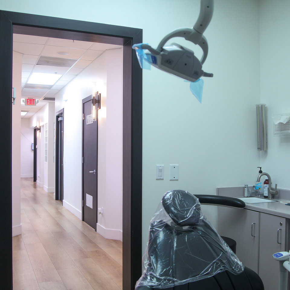 Dental office room with a black chair covered in plastic, overhead light, and view of a hallway.