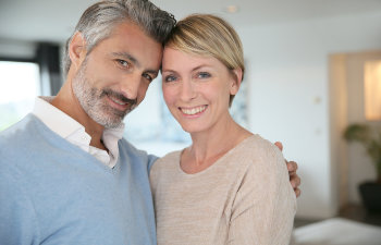 A smiling couple poses together indoors, with the man wearing a light blue sweater and the woman in a beige top.