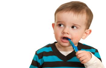 A toddler in a striped shirt holds a toothbrush in their mouth against a plain white background.