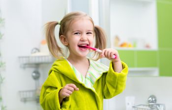 A young child in a green robe is brushing their teeth with a pink toothbrush, smiling in a bathroom setting.