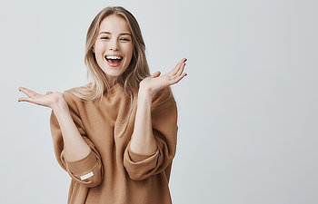 Woman in a brown sweater smiling with hands raised against a light gray background.