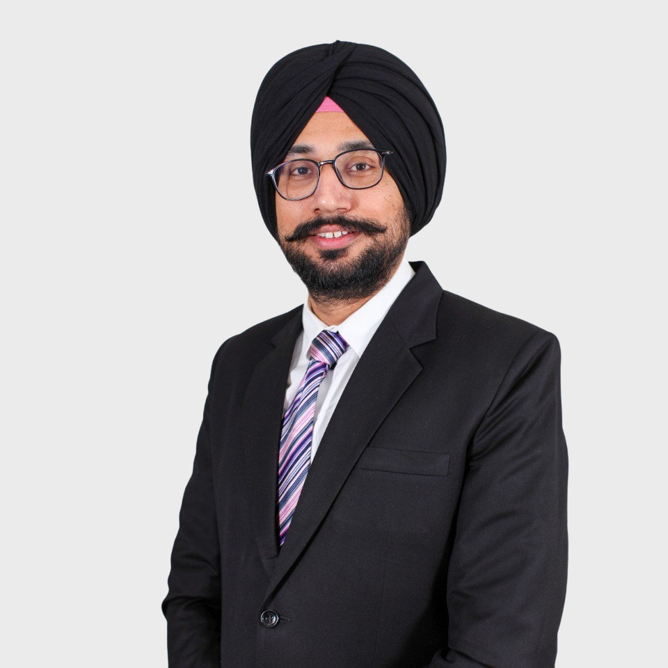 Person wearing a black turban, glasses, and a black suit with a striped tie, smiling against a plain background.