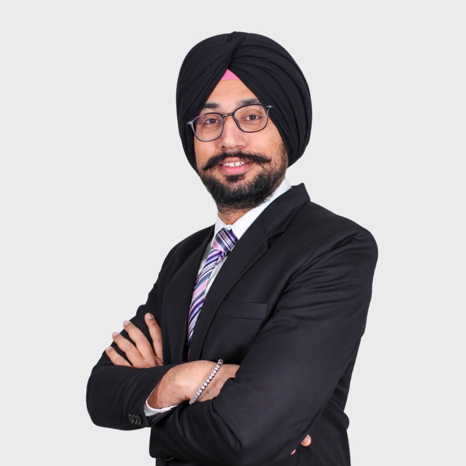 Person wearing a suit and turban, with arms crossed, smiling, against a plain background.