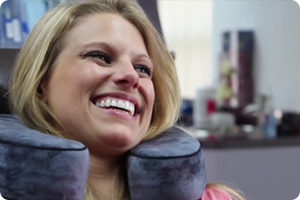 A woman smiles while wearing a gray neck pillow, seated indoors with blurred background.