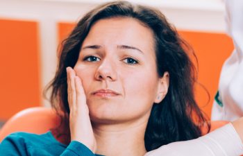 A woman with a concerned expression holds her cheek, sitting in a medical setting.