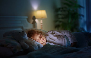 Child sleeping peacefully in bed at night, with a soft glowing lamp in the background and a stuffed animal beside them.