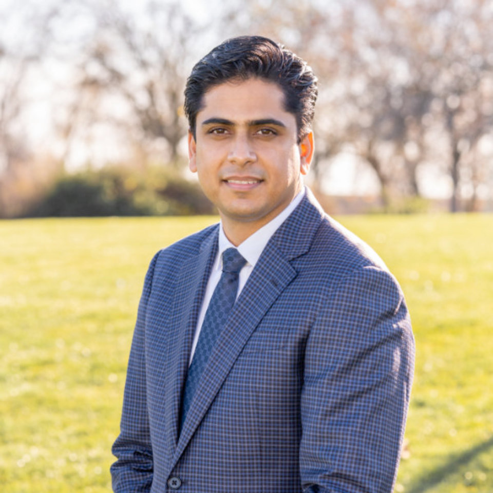 Person in a blue checkered suit and tie stands outdoors on a sunny day, with trees and grass in the background.