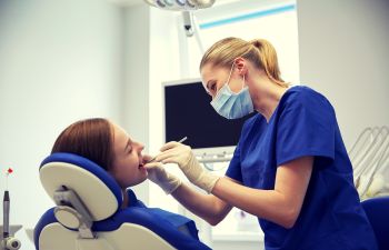 Dentist in blue scrubs examines a patient's mouth with dental tools in a clinic room.