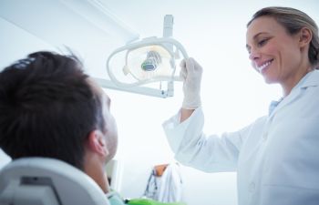 A dentist wearing gloves adjusts an overhead light while smiling at a patient sitting in a dental chair.