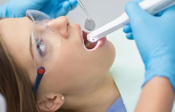 Person receiving a dental examination, wearing safety glasses. A dentist uses a mirror and a dental instrument while wearing blue gloves.