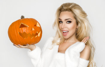 Person smiling while holding a carved jack-o'-lantern, wearing a white outfit against a plain background.