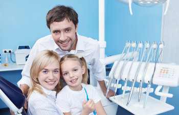 A woman, man, and a young girl with a toothbrush smile at the camera in a dental office. Dental equipment is visible in the background.