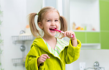 A young child in a green robe is brushing her teeth with a pink toothbrush, smiling in a bathroom.