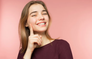 Smiling person with long hair points to their cheek against a pink background, wearing a dark top.