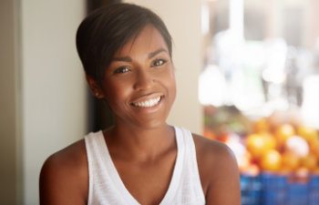 A person in a white tank top smiles at the camera. In the blurred background, there are baskets of oranges.