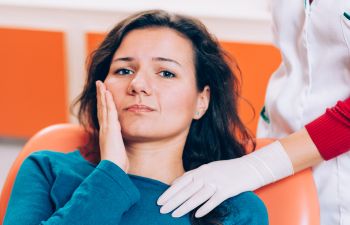 Woman sitting in a dental chair, holding her cheek with a pained expression while a gloved healthcare professional stands beside her.