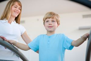 A woman smiles at a child in a light blue shirt who is standing and holding onto rails indoors.