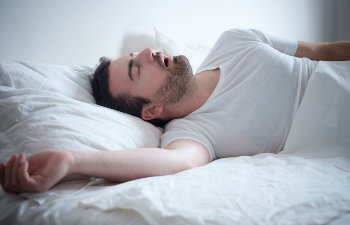 A man with a beard is sleeping on his back in bed, mouth open, wearing a white T-shirt.