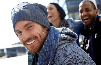 A man in a beanie and sweater smiles at the camera. Two people in the background are also smiling and laughing.