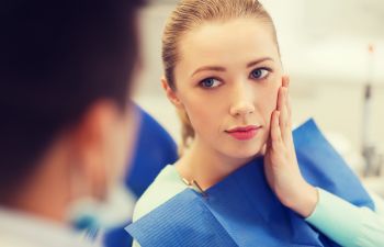 Woman sitting in a dental chair with a blue bib, touching her cheek, looking at a person in the foreground.