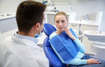 A woman sits in a dental chair touching her cheek, talking to a dentist.