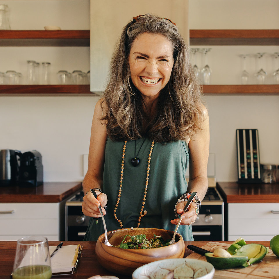 A person smiles while preparing a salad in a kitchen. A glass, notebook, and other kitchen items are visible in the background.