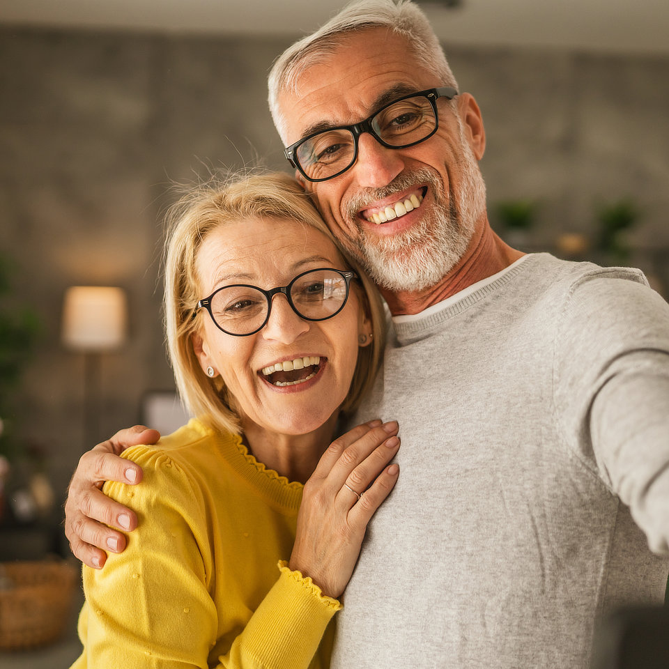 Older couple wearing glasses, smiling and embracing while taking a selfie indoors. The man has gray hair and a beard and wears a gray sweater. The woman has blonde hair and wears a yellow top.