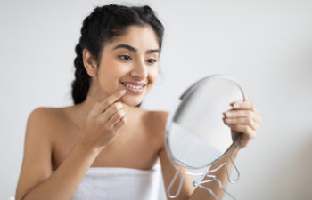 A woman wrapped in a towel looks into a handheld mirror, smiling and touching her chin.