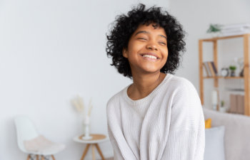 A person with curly hair smiles while wearing a light sweater, sitting in a bright room with shelves and decor in the background.