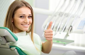 Person sitting in a dental chair, smiling, and giving a thumbs-up. Dental equipment is visible in the background.