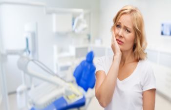 Woman in a white shirt holding her cheek, looking uncomfortable, standing in a dental office.