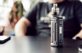 A vape device sits on a table with a blurred person in the background.