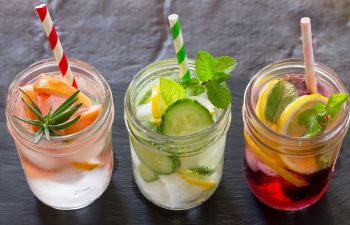 Three glass jars filled with iced drinks: one with grapefruit and rosemary, one with cucumber and mint, and one with lemon slices and a red liquid, each with a striped straw.